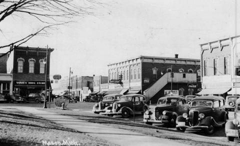 Black and white photo of street scene in Mason, Michigan, with parked cars in the foreground. An intersection takes up the middle ground. Buildings distinguished by tall second stories and high windows, with cars parked below them, make up the background.