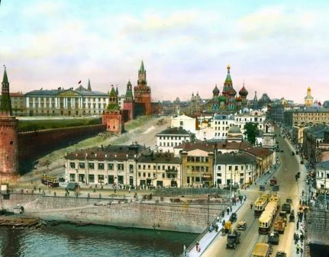 A vintage color photo from the 1930s shows a busy Moscow bridge on a clear day. The bridge is in front of a greenish onion-domed church in the center-background and the red towers of the Kremlin fortress along the left side of the image.
