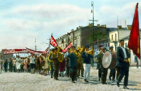 Colorful sunlit photo shows a worker parade on a street in Moscow in 1931. The parade is led by an all-male brass band, with a drummer boy in the lead; behind the band are women carrying a flowing red banner with large white lettering.