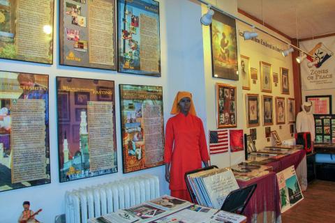 Contemporary color photo of exhibits at the Muslim American Museum & Archive. A mannequin of a dark-skinned young Muslim woman of African descent is shown wearing a red muslin dress and orange hajib. She stands directly in front of a blue-painted wall filled with colorful images and commentary on traditional beliefs, practices, and values of Islam.