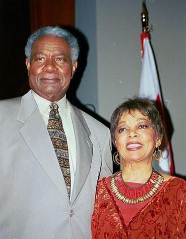 Color photo. Tall, muscular, gray-haired Ossie Davis stands next to petite, radiant Ruby Dee. Davis wears a light gray suit and colorful African Kente tie. Dee wears an elegant red dress with interwoven figures, gold earrings, and a wide African style necklace.