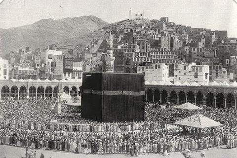 Vintage black and white photo of the Kaaba and Grand Mosque, in Mecca. The exterior of the Kaaba is covered with a large black cloth. It stands in the center of the photo, in front of the Grand Mosque, in the near background. In the far background is a mountain with numerous buildings.]