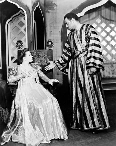 Black and white photo from the Theater Guild’s production of Othello. Paul Robeson’s character Othello is standing at right, with an extended right hand, casting an angry look at Uta Hagen’s seated Desdemona at left. Robeson is dressed in a satin striped robe, and Hagen is wearing a white silk gown.