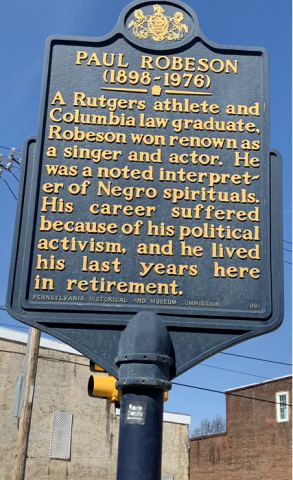 A color photo of a historical marker plaque, yellow letters against dark blue, in front of the Paul Robeson House and Museum, near the intersection of 49TH and WALNUT ST. The plaque reads: PAUL ROBESON (1898-1976)/A Rutgers athlete and Columbia law graduate, Robeson won renown as a singer and actor. He was a noted interpreter of Negro spirituals. His career suffered because of his political activism, and he lived his last years here in retirement. - Pennsylvania Historical and Museum Commission 1991.