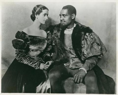 Black and white photo shows Paul Robeson and Peggy Ashcroft seated on stage in period-piece costumes for their roles in the 1930 production of Othello at London’s Savoy Theatre.
