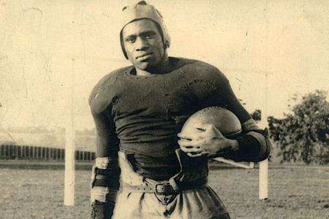 Black and white photo of Paul Robeson, posed standing in his football uniform, wearing a leather helmet and cradling a football in his left arm.
