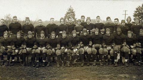 Black and white photo of the 1918 Rutgers football team, posed in their uniforms and leather helmets. The front row is seated, the back row is standing. Paul Robeson is seated near the end of the front row.