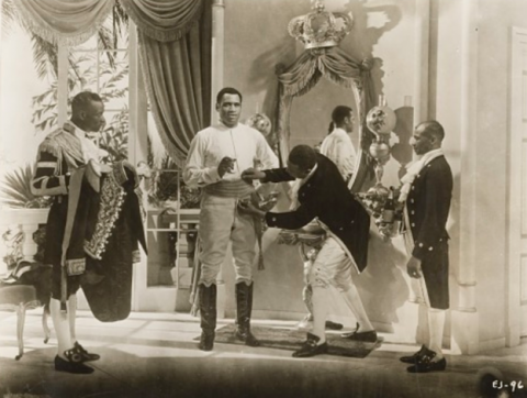 A black and white photo from a scene in the 1933 film Emperor Jones, showing Paul Robeson’s character being dressed in his military uniform by several costumed Black servants.
