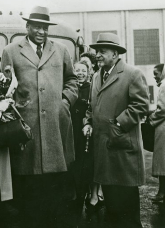 Black and white photo from 1949. Paul Robeson stands in the foreground beside W.E.B. Du Bois. A smiling young woman and what appears to be a small bus appear in the middle ground. Robeson is markedly taller than Du Bois. Both men are wearing gray-toned wool suits and fedoras.