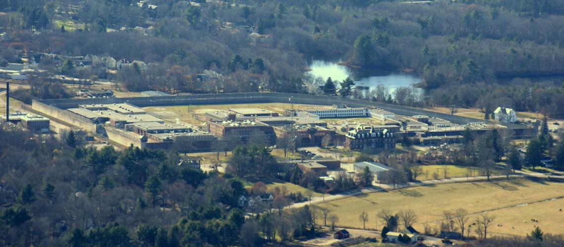 Color photo highlights the wall-enclosed prison facility, shown in the left-to-right center of the image, in its woodland location, with a river flowing just beyond the rear wall. The rest of the photo shows some unidentifiable low buildings enclosed by woods.