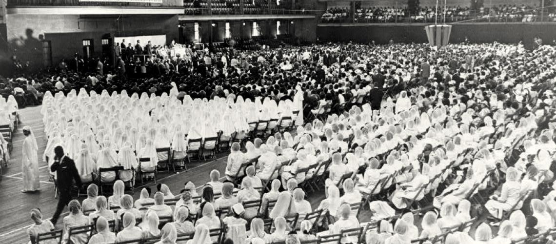 Black and white photo from 1965, a massive indoor venue with thousands of Nation of Islam followers. The foreground to center of the photo shows several hundred women wearing white abayas, seated in folding chairs. A banner hung in the far-left balcony reads THERE IS NO GOD BUT ALLAH AND MUHAMMAD IS HIS PROPHET.