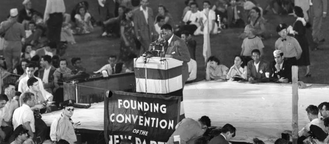 Black and white photo. Paul Robeson stands at microphones that are positioned on a podium on a raised stage. A banner that reads PROGESSIVE PARTY NATIONAL CONVENTION hangs from a wire. People below the stage are moving about doing various things. A policeman wearing sunglasses stands near the stage.
