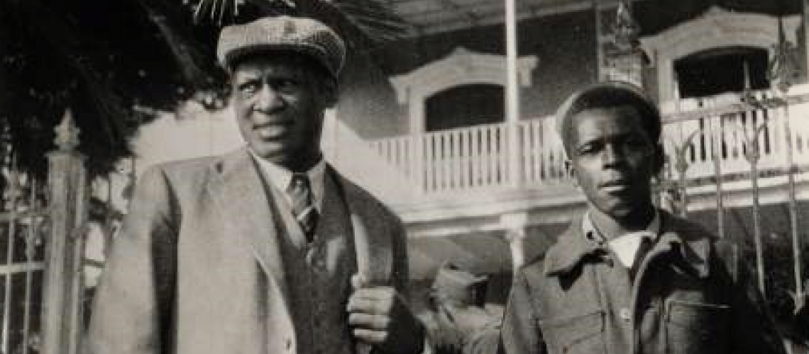 Black and white photo of Paul Robeson, shown in the foreground standing beneath a palm tree beside a young African American man who is a volunteer with the International Brigade in the Spanish Civil War. Robeson wears a light-colored suit with vest, striped tie, and English cap. The volunteer wears a dark, buttoned jacket. In the background is a Spanish colonial-style building with a white-wood balcony railing.
