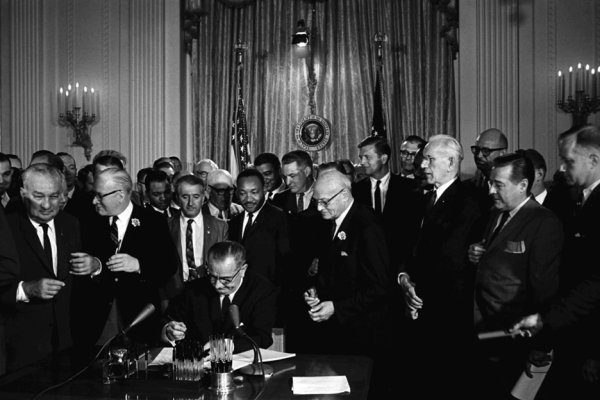 Black and white photo shows a seated President Lyndon B. Johnson surrounded by male dignitaries, most of whom are white, as he signs the 1964 Civil Rights Act. Dr. Martin Luther King Jr., one of the few Black people in the photo, stands directly behind the president to view the signing.