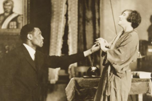 Black and white photo of Paul Robeson performing on stage with an unidentified actress in the 1924 production of Eugene O’Neil’s play All God’s Chilluns Got Wings. Robeson’s hand is extended to touch the actress’s cupped hands. The setting is an indoor room.