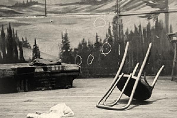 Black and white photo of the stage in the aftermath of Malcolm’s assassination. An upturned chair is in the foreground, the wall of the stage, with several bullet holes marked by chalked circles in the background.