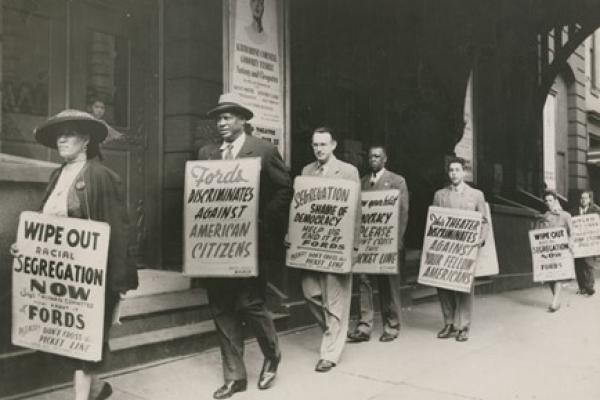 Black and white photo. Paul Robeson shown marching in front of the theater. Like others in the demonstration, he carries a protest sign hung from his shoulders. His sign reads FORDS DISCRIMINATES AGAINST AMERICAN CITIZENS. Robeson appears behind a woman in an oval hat carrying a sign protesting racial segregation at Fords Theater; he wears a dark suit and a fedora.