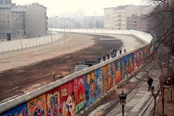 Color photo of a section of the Berlin Wall in the winter of 1986. This image shows the colorful murals of the outer wall, painted by West Germans on their side of the border. The infamous Death Strip appears below and beyond the outer wall. The Death Strip meets an interior wall that provides a barrier to potential fugitives. Bleak, gray, shuttered apartment buildings hover over the interior wall.