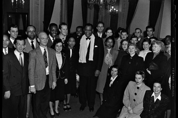 Black and white photo. A smiling Paul Robeson stands beneath a candelabra surrounded by a racially integrated group of men and women at the California Labor School. He wears a black tuxedo with black tie, and a scarf around his neck.