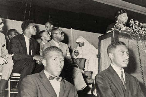 Black and white photo. Cassius Clay, soon to adopt the name Muhammad Ali, is seated behind two Fruit of Islam guards. He looks toward the speaker podium where Elijah Muhammad addresses an audience. Elijah Muhammad wears a male Muslim prayer hat. Clay wears a dark suit, white shirt, and dark tie, without a hat.