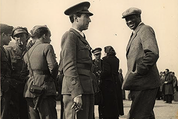 Black and white photo from 1938 showing, in the foreground, a smiling Paul Robeson, dressed in a suit, with his left hand in a pocket, wearing an English wool cap; and Robeson’s Spanish guide, Captain Fernando Castillo, dressed in a military uniform and hat, with his right hand on a walking stick.