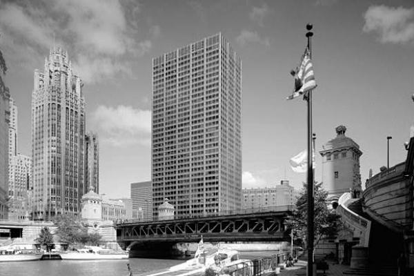 Black and white photo of tall buildings in Chicago. There is a flagpole flying an American flag, a river with some boats tied up along its edge. A staircase leads up to a steel bridge that crosses the river.