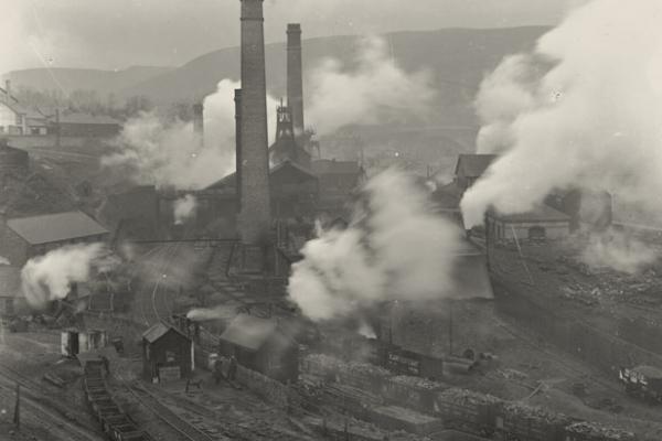A black and white photo of the Cymmer Colliery in the coal-mining village of Porth, in the Rhonnda Valley of Wales, around 1905. Smoke surrounds the towered buildings of the colliery, which appear against a background of low mountains and a rain-laden sky.