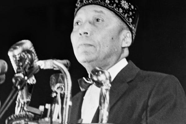 Black and white photo of Elijah Muhammad standing behind a podium with several microphones. He is wearing a white shirt with a bow tie, a dark suit jacket, and an embroidered dark brimless hat. He is cleanshaven.