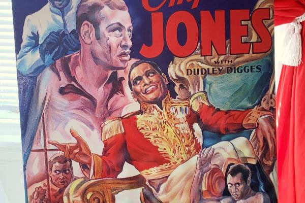 A colorful poster advertises the 1933 film Emperor Jones. The poster displays five images of Robeson at the various stages of his character’s rise as a corrupt dictator. Four of the images revolve around Robson dressed in a red and gold military uniform, with gold shoulder epaulettes evocative of the era of Napoleon Bonaparte.