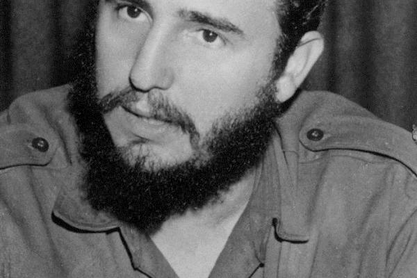 Black and white photo of Fidel Castro with dark hair and beard wearing fatigues.