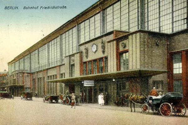 A color postcard from the mid-1920s of the Berlin Friedrichstrasse train station, with horse-drawn black carriages stationed on the street in front of the building.