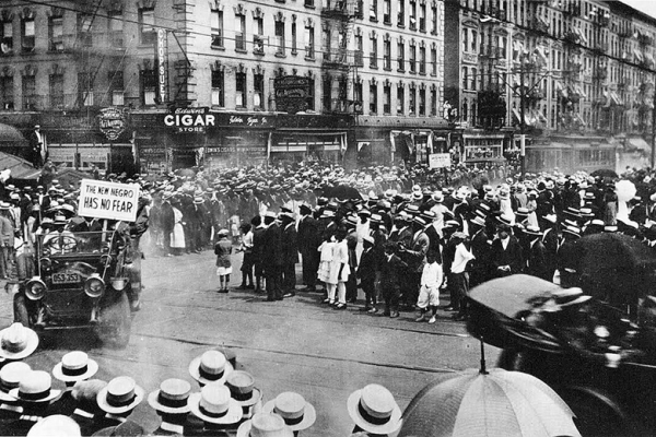 Black and white photo of the parade as it reaches an intersection in Harlem. Large crowds line the street. Parade participants ride in open cars in the foreground. A car shown turning at the intersection carries a plaque that reads THE NEW NEGRO HAS NO FEAR. A row of four-story tenement buildings, covered with fire escapes, form the background of the photo; a large white cigar sign appears on a tenement near the intersection.