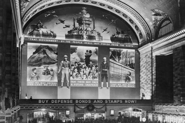 Black and white photo of Grand Central station mural honoring U.S. Navy and U.S. Army in the wartime 1940s. The signage reads BUY DEFENSE BONDS STAMPS NOW! Scenes illustrate what war bonds supported. Images of a sailor and an infantryman are positioned between the scenes. The top of the mural features a tank on both sides of a large battleship with airplanes in the sky behind them, and is framed under the curve of the station’s dome ceiling.
