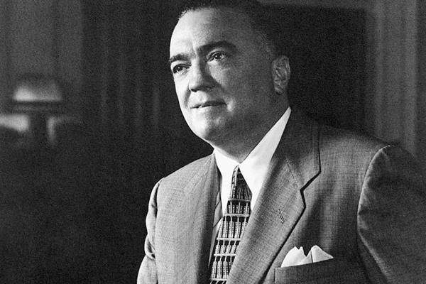 Alt text: Black and white photo of J. Edgar Hoover wearing a patterned tie, white shirt, mid-tone suit jacket with a white pocket handkerchief