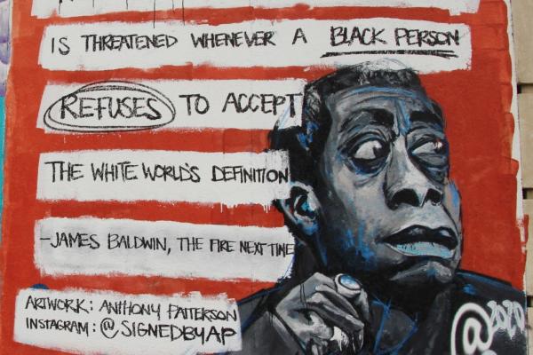 Contemporary color photo of a mural of James Baldwin. The mural features an image of Baldwin, which is painted gray with blue highlights and depicts the activist in a reflective pose. A quote from Baldwin’s THE FIRE NEXT TIME is hand-printed in black letters, the words distributed in several white bubbles arrayed against a red backdrop that frames the entire mural. The quote is: THE POWER OF THE WHITE WORLD IS THREATENED WHENEVER THE BLACK WORLD REFUSES TO ACCEPT THE WHITE WORLD’S DEFINITION.