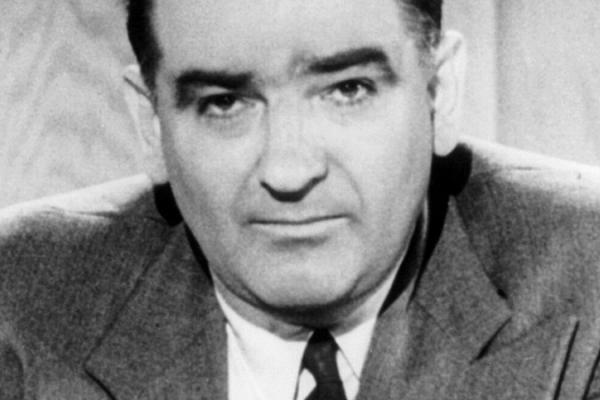 Black and white iconic photo portrait of Senator Joseph McCarthy in 1954. McCarthy is distinguished by his v-shaped hair line. He has a bulbous nose and large ears. He is shown wearing a gray-toned wool suit jacket with large lapels and a dark tie.