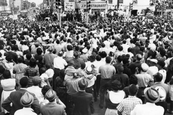 Black and white photo showing a large crowd of men, women, and children in the intersection at 40th and Market St., congregating for a rally led by Dr. Martin Luther King Jr.  Dr. King and other Black leaders stand on a platform in front of the JP RESTAURANT, on the north side of the intersection.