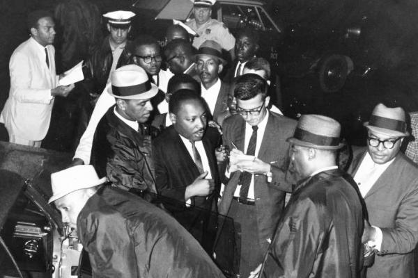 Black and white photo showing Dr. Martin Luther King Jr. surrounded by a group of Black male colleagues on his arrival at Philadelphia International Airport. Several of the men wear fedoras. Dr. King wears a dark suit and light gray-tone tie. He is talking with a white reporter, who is taking notes. Dr. King’s close associate Andrew Young is attired in a light suit and dark tie and appears in the left background.