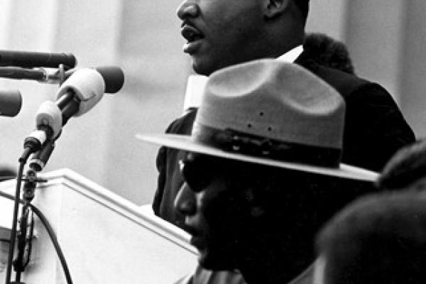 Black and white photo of Dr. Martin Luther King, Jr. speaking into a microphone with a state trooper near him