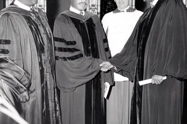 Black and white photo of Dr. Martin Luther King Jr. standing between two Black pastors. Dr. King and Rev. Shepard Sr. wear dark academic gowns. Rev. Shepard Jr. wears a traditional black ministerial robe; he and Dr. King shake hands. A fourth person is a white priest, robed in a white gown with a dark clerical collar. Stain-glass windows form the backdrop of the photo.