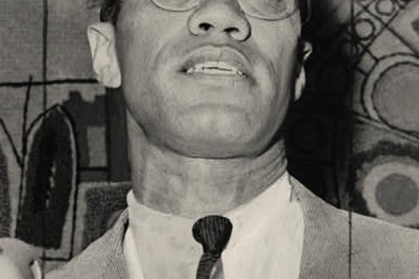 black and white photo of Malcolm X, at three microphones, head tilted upward, clean-shaven with closely cropped hair and eyeglasses, wearing a light gray-toned suit or sports coat, white shirt, and dark tie.