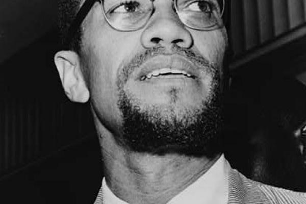 Black and white photo of Malcolm X in 1964. He is standing, wearing a light corduroy jacket, a white shirt, and a dark tie. His head is tilted to his left side. Emblematic of his break with the Nation of Islam, he now has a trimmed moustache connected to a small goatee, forming a circle.