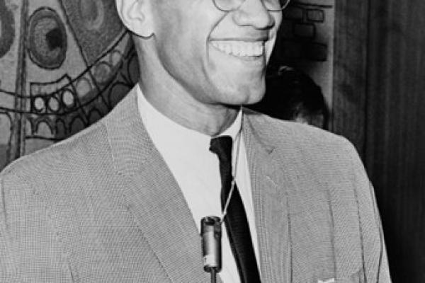 Black and white photo of Malcolm X in 1964. Shown smiling and relaxed, he stands at a microphone. He wears a light gray-tone suit or sports coat, with a dark tie. He has closely cropped hair, is clean shaven, and wears glasses.