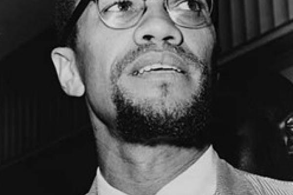 Black and white photo of Malcolm X in 1964. He is standing, wearing a light corduroy jacket, a white shirt, and a dark tie. His head is tilted to his left side. Emblematic of his break with the Nation of Islam, he now has a trimmed moustache connected to a small goatee, forming a circle.