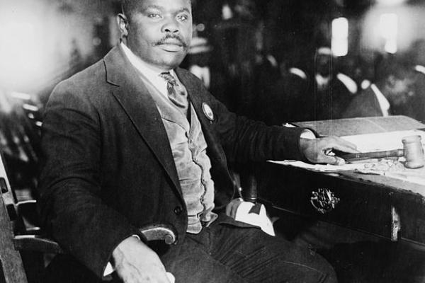 alt text=Black and white photo of Marcus Garvey. He is seated and his chair is turned away from the desk to face the camera. He is dressed in a dark suit, with a buttoned light gray-tone vest and striped tie.