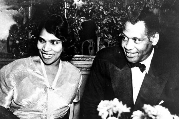 Black and white photo of Paul Robeson and Marian Anderson, seated at a table decorated with a flower arrangement. Robeson, at right in the photo, wears a dark tuxedo with black tie; Anderson’s torso is covered by a formal white blouse. Both Robeson and Anderson are smiling.