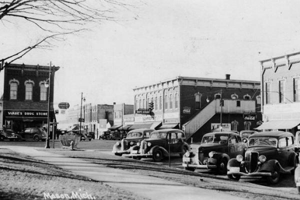 Black and white photo of street scene in Mason, Michigan, with parked cars in the foreground. An intersection takes up the middle ground. Buildings distinguished by tall second stories and high windows, with cars parked below them, make up the background.