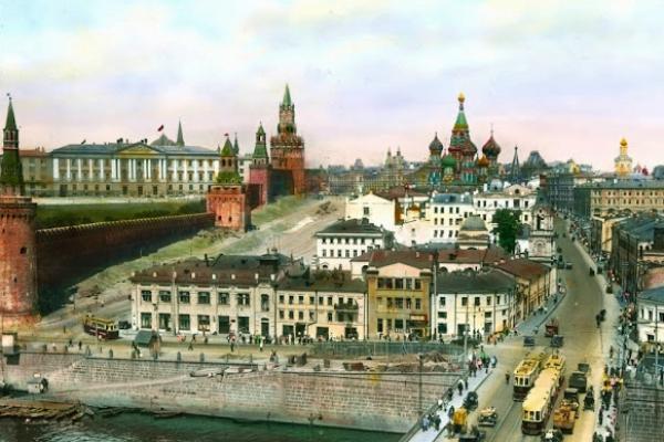 A vintage color photo from the 1930s shows a busy Moscow bridge on a clear day. The bridge is in front of a greenish onion-domed church in the center-background and the red towers of the Kremlin fortress along the left side of the image.