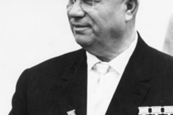 Black and white photo of Nikita Khrushchev in 1963. Khrushchev is distinguished by his bald head and bulbous face. He wears a dark suit and dark tie; a row of medals appears chest-high on the right side of his jacket. He holds assorted papers in his hands.