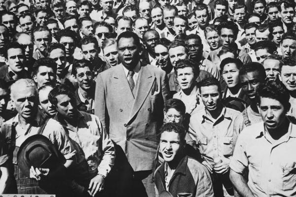 Black and white photo. A singing Paul Robeson stands tall in the foreground of this photo, surrounded by a crowd of all-male shipyard workers. Robeson wears a double-breasted, light-colored wool suit; both he and the shipyard workers are hatless.